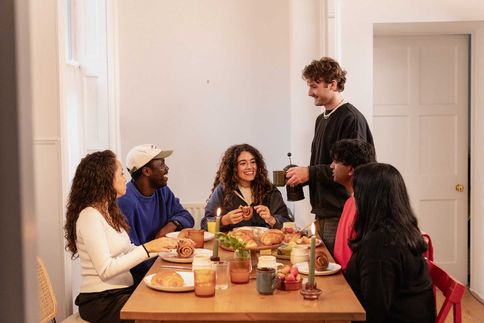 Six friends around a table over coffee and pastries, one of them standing to pour